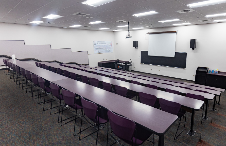 Tiered classroom with long rows of tables and purple chairs arranged stadium-style, facing a projection screen, chalkboard, wall-mounted speakers, and instructor podium at the front.