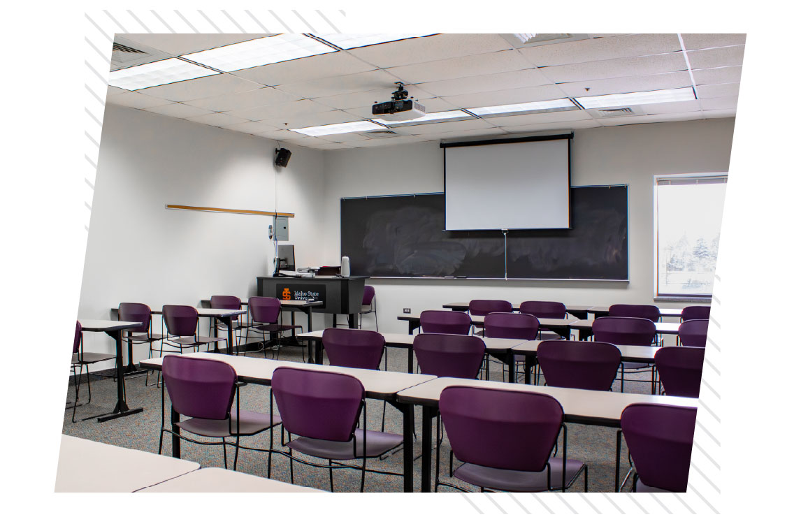 HE classroom with rows of tables and purple chairs facing a teaching station, chalkboards, and a ceiling-mounted projector with a projection screen.