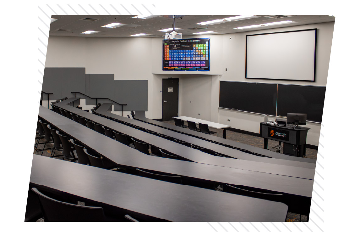 Large tiered classroom at ISU Idaho Falls with rows of fixed tables and chairs facing blackboards, a projection screen, instructor station, and periodic table display.
