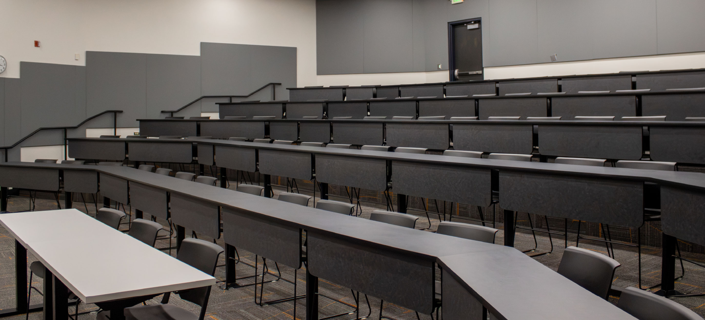 Side view of the CHE Large Tiered Classroom at ISU Idaho Falls, showing multiple rows of fixed gray tables and chairs arranged on tiered levels with stair access along the walls.