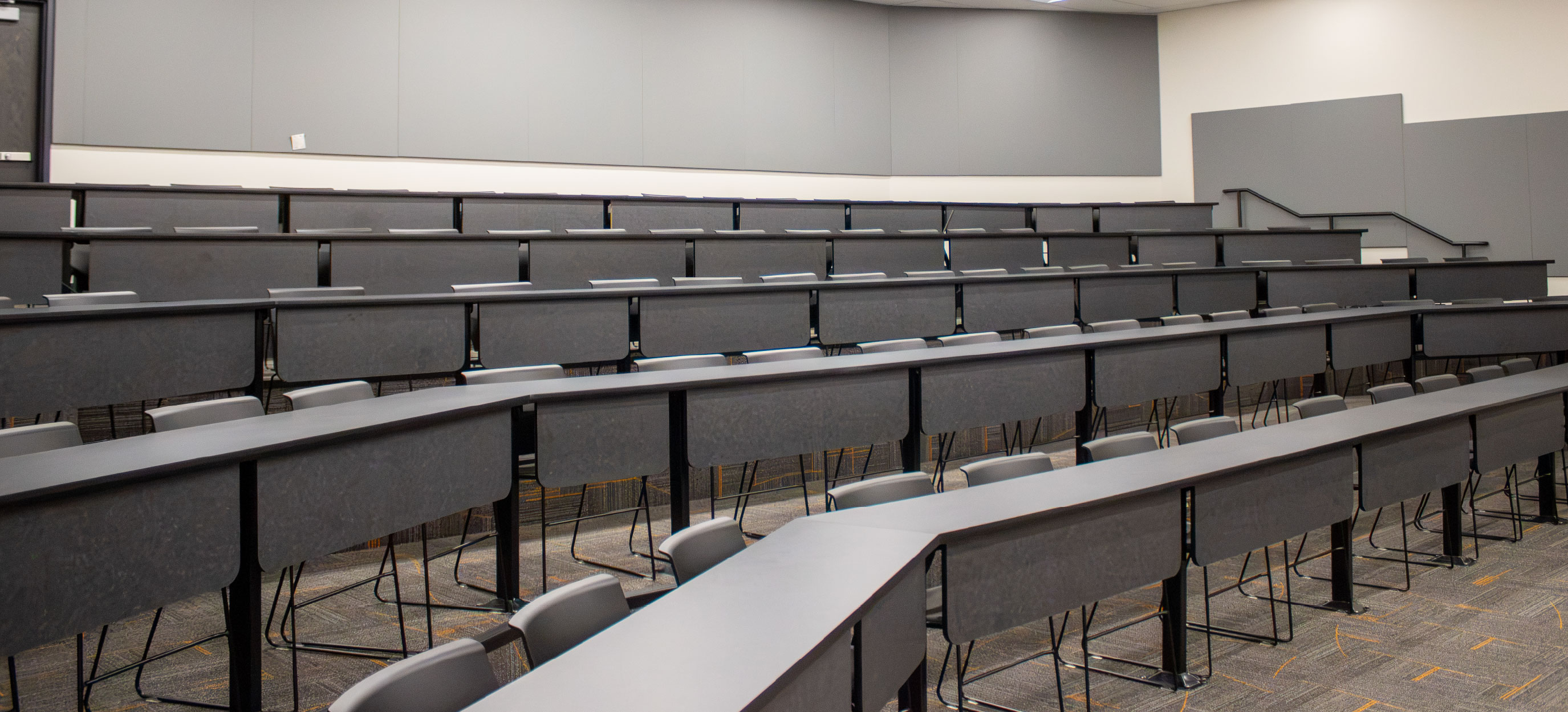 Rear view of the CHE Large Tiered Classroom at ISU Idaho Falls, showing multiple rows of fixed gray tables and chairs arranged on tiered levels with stairs along the side.