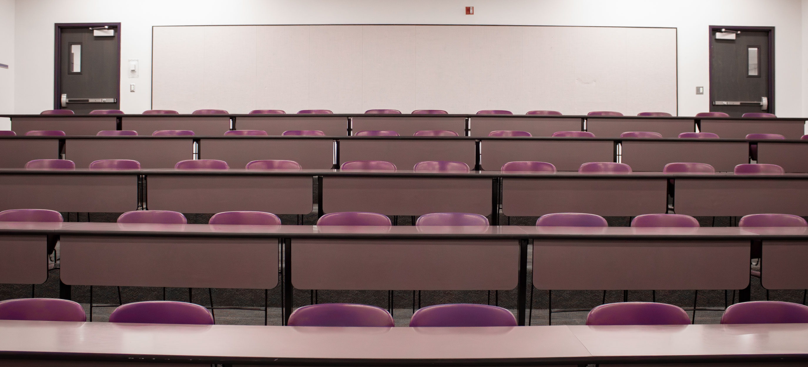 Rear-facing view of the CHE Tiered Classroom at ISU Idaho Falls, showing fixed rows of tables and purple chairs with a large whiteboard and two exit doors at the back of the room.