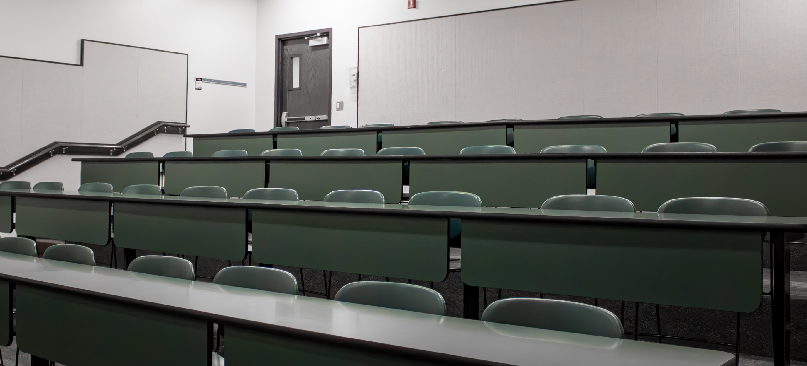 Small CHE tiered classroom with fixed rows of green desks and chairs arranged on ascending levels, with stairs and handrails along the side.