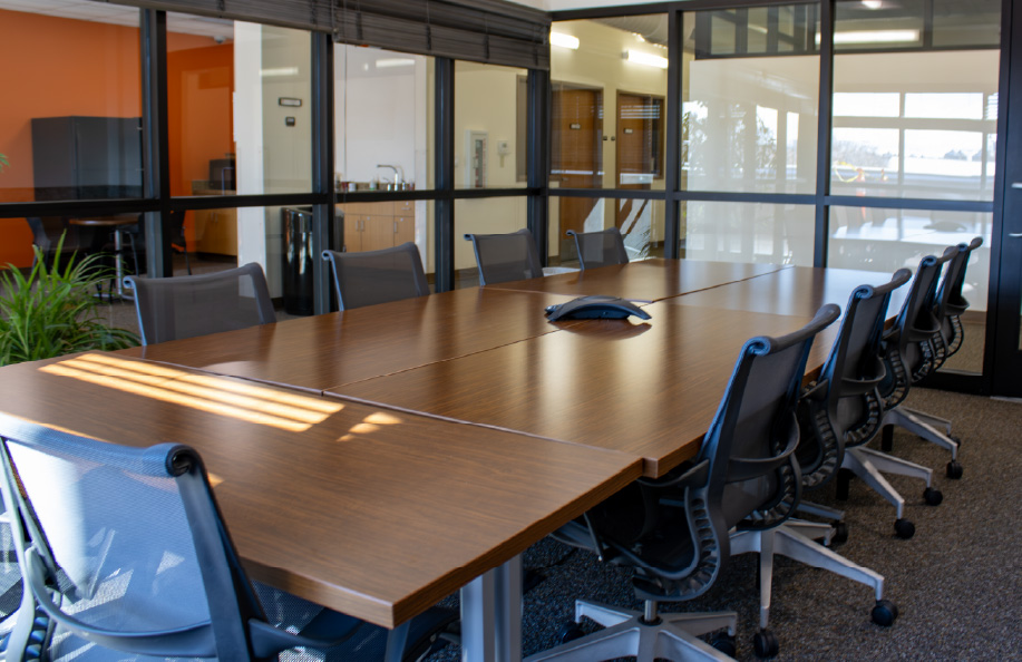 Modern conference room with a long wooden table and mesh office chairs, enclosed by glass walls, with a conference phone centered on the table and natural light coming through nearby windows.