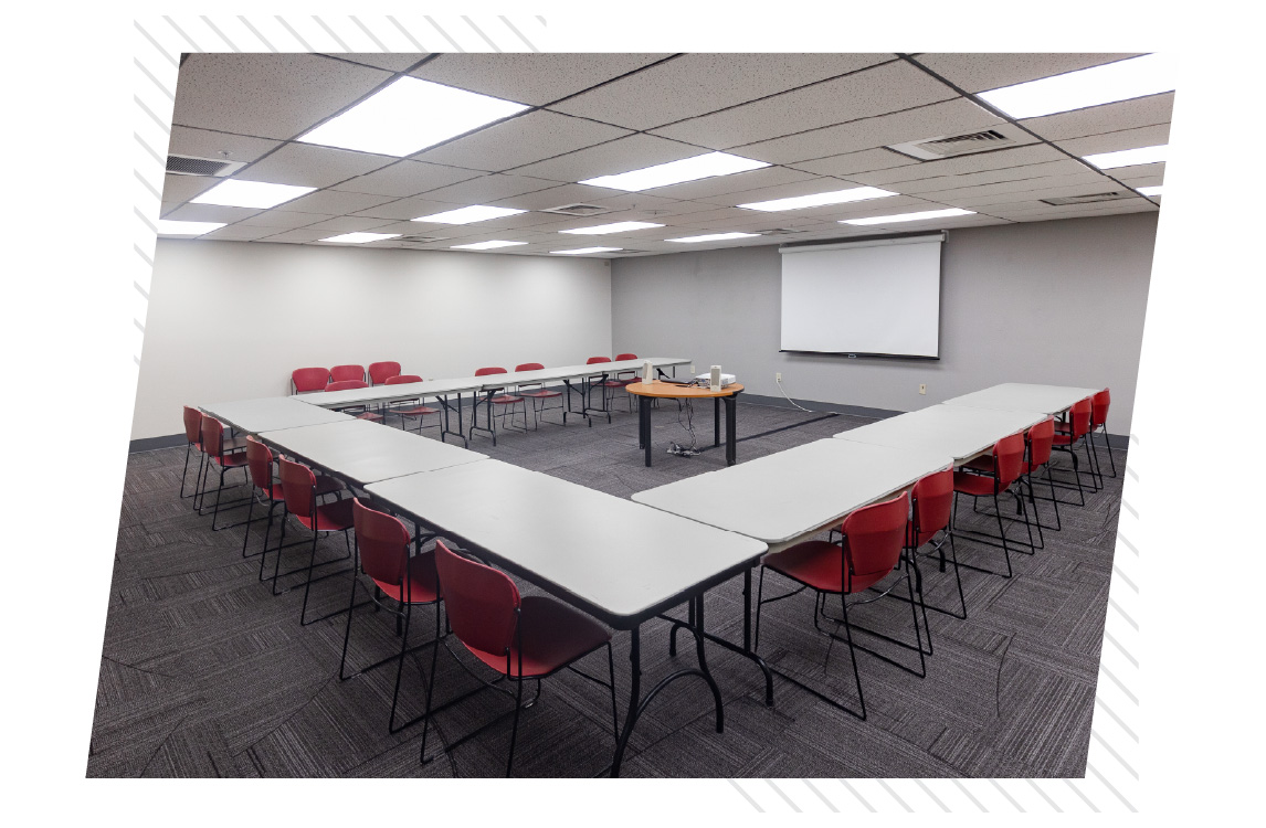 Spacious meeting room set up in a U-shape configuration with white tables and red chairs, facing a projection screen and small presentation table at the front.