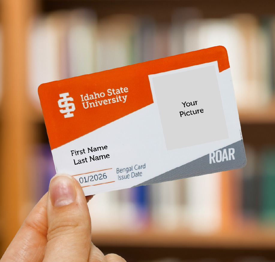 A close-up photograph of a hand holding an Idaho State University Bengal ID card template. The hand's thumb and index finger hold the bottom-left corner of the card, which is angled slightly forward.