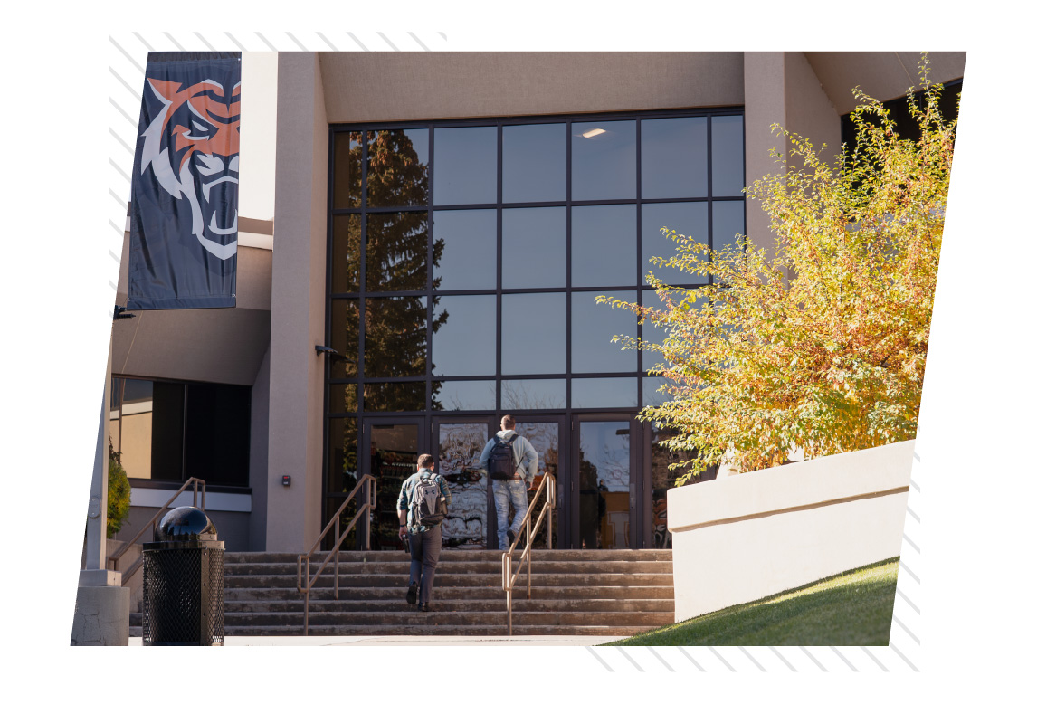 Two students with backpacks walk up the stairs and enter a glass-fronted building on the Idaho State University Idaho Falls campus, with a Bengal banner hanging beside the entrance and a tree with fall-colored leaves nearby.