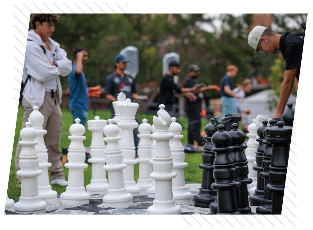 Students gather around a giant outdoor chess set, with oversized black and white chess pieces in the foreground and students socializing in the background.