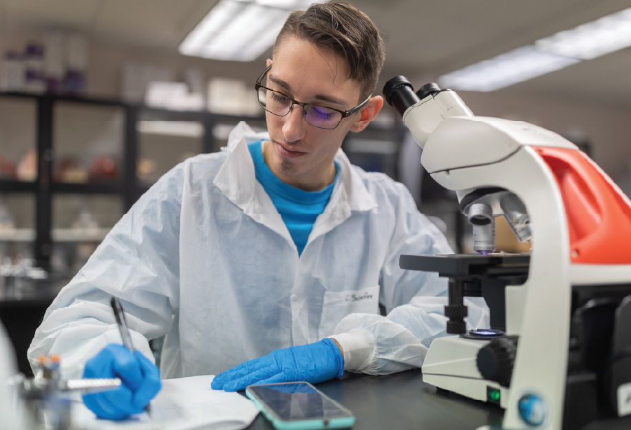 Student wearing a white lab coat, blue gloves, and safety glasses writes notes at a laboratory workstation beside a microscope.