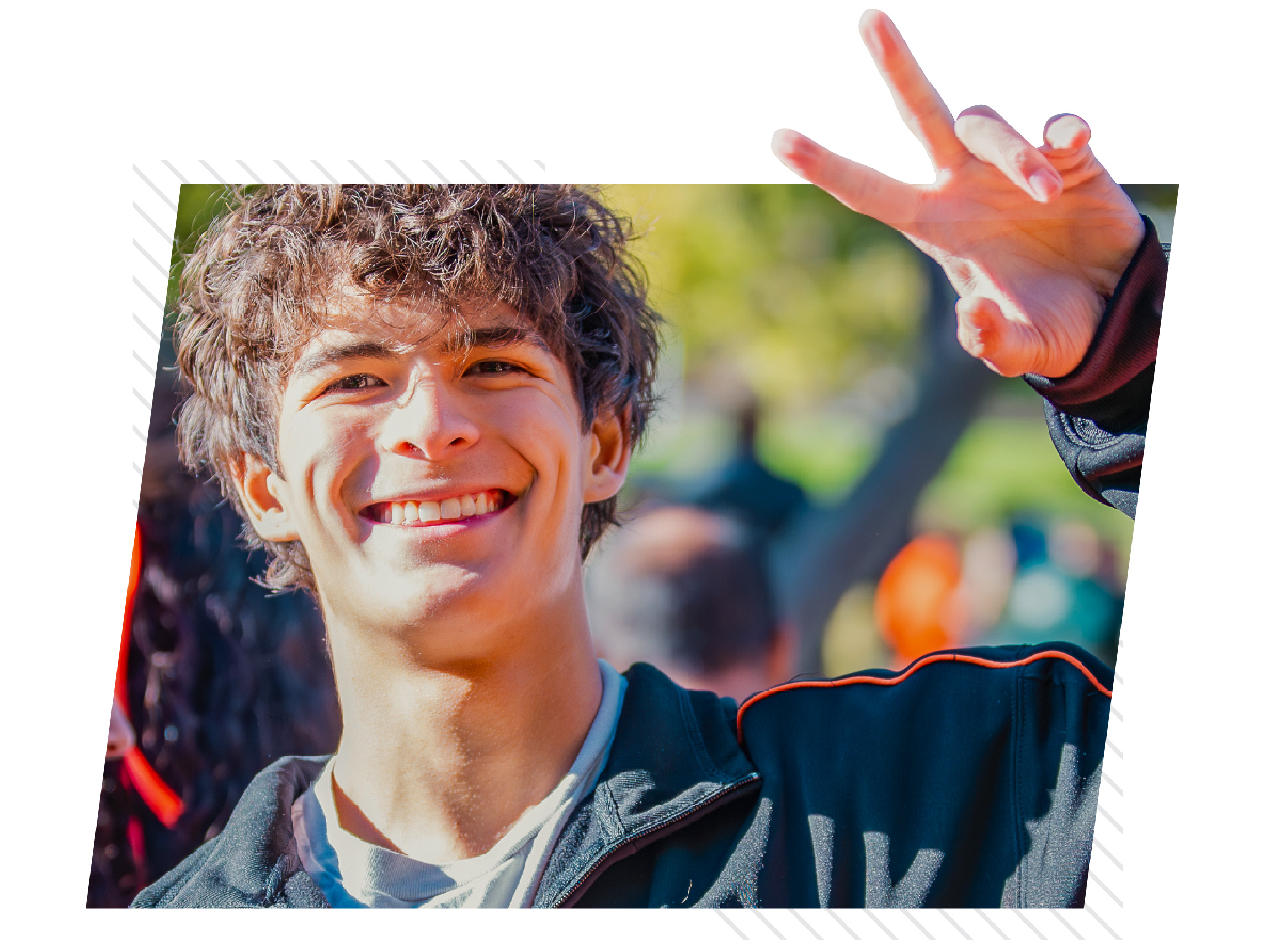 Close-up of a smiling college student outdoors making a peace sign with one hand. Trees and other students are softly blurred in the background, creating a lively and welcoming campus involvement atmosphere.