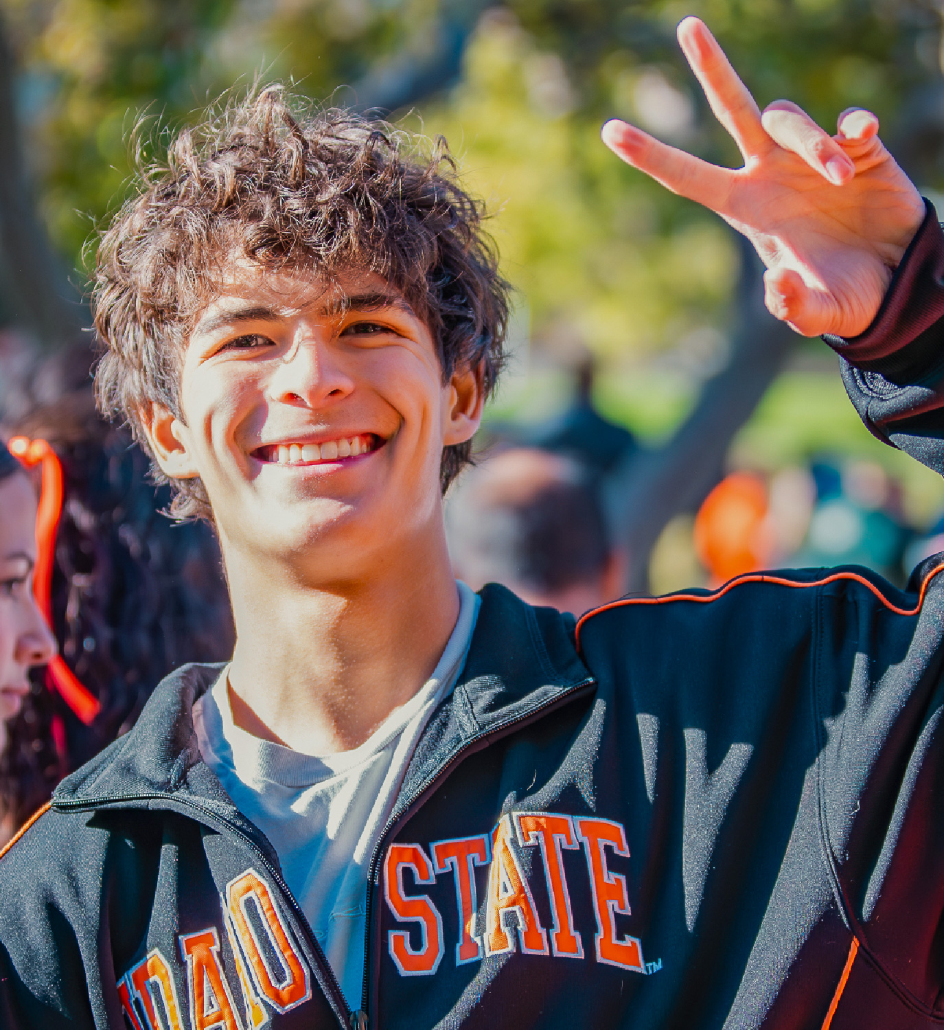Close-up of a smiling college student outdoors wearing a black Idaho State jacket with orange lettering. The student raises one hand in a peace sign, with trees and other students softly blurred in the background, creating a lively campus atmosphere.