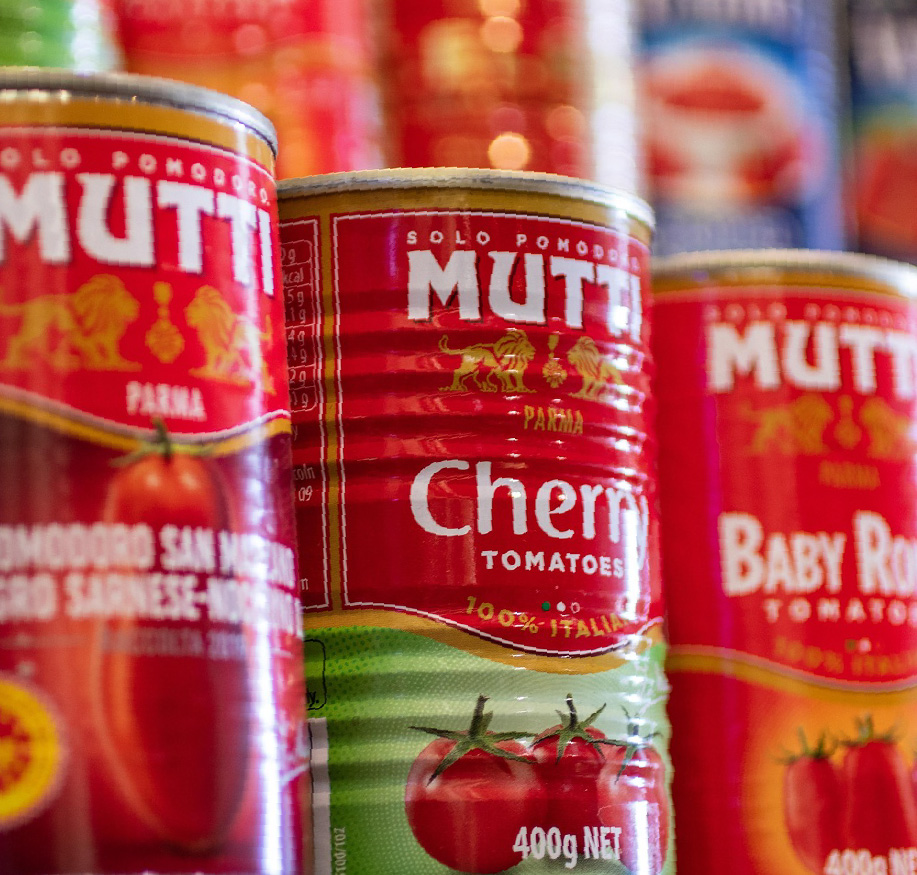 Close-up of several red cans of Mutti tomatoes displayed on a shelf. The labels show different varieties, including cherry and baby Roma tomatoes, with colorful packaging and tomato images on the front.
