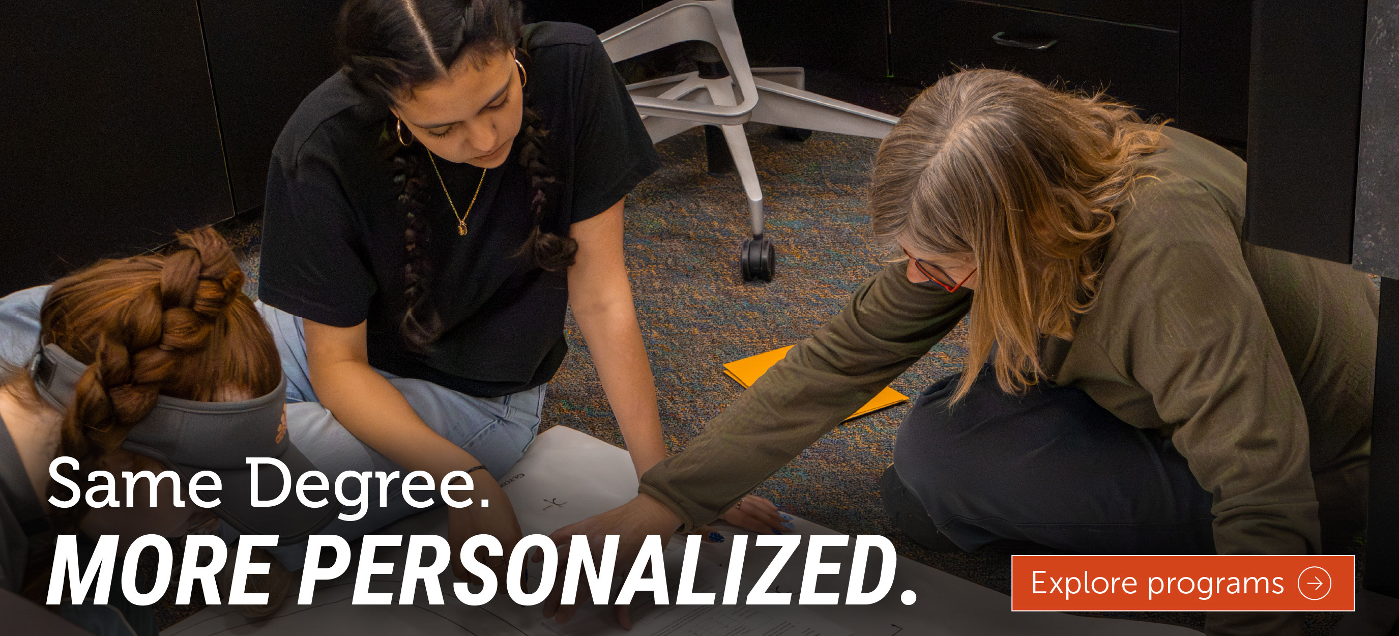 A professor works one-on-one with two students as they review materials spread across the floor during a collaborative learning activity. The image includes the text, 