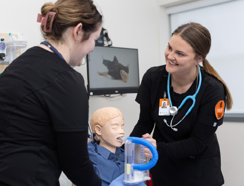 Two students in a healthcare classroom practice a medical procedure using a training mannequin. One student holds a breathing device while the other assists, with medical equipment visible in the room.