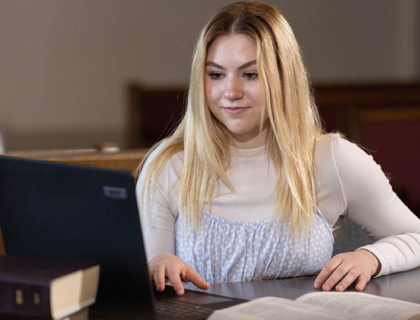 Student sits at a table using a laptop while reading from an open book in a quiet study space.