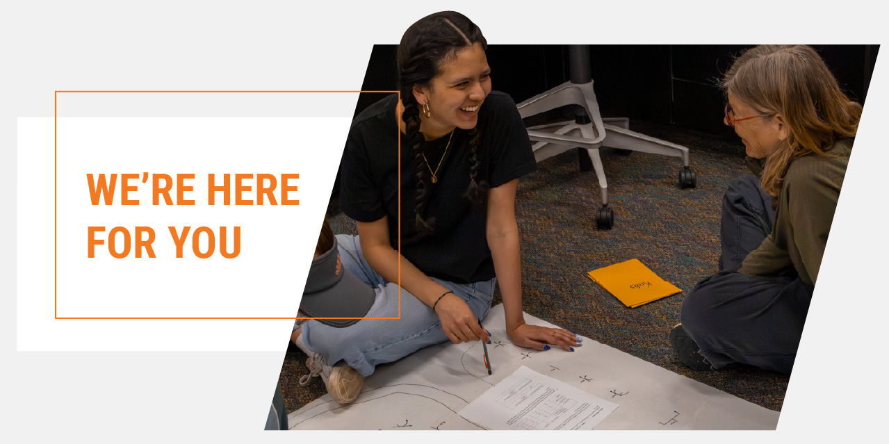 A student and an advisor sit on the floor together, smiling as they review papers and collaborate on an activity. The image includes the text, 