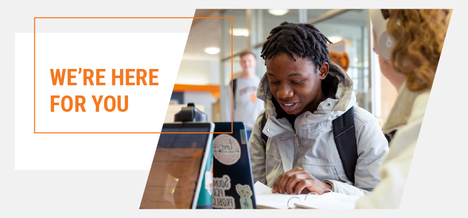 Student wearing a backpack reviews paperwork at a campus service desk while speaking with a staff member; on-screen text reads ‘We’re here for you.’