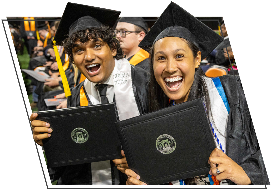 Two smiling Idaho State University graduates in black caps and gowns hold their diploma covers and celebrate during commencement. The graduates face the camera with excited expressions while seated among fellow graduates, with orange graduation accents visible in the background. The image is framed with an angled black border on a white background.