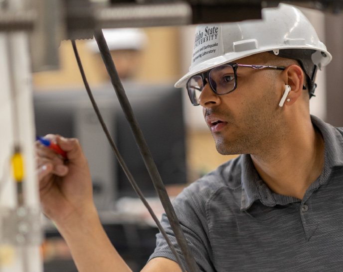 A student wearing a white Idaho State University hard hat and safety glasses marks measurements on industrial equipment in an engineering laboratory. The student focuses on the task while working in a hands-on technical learning environment.