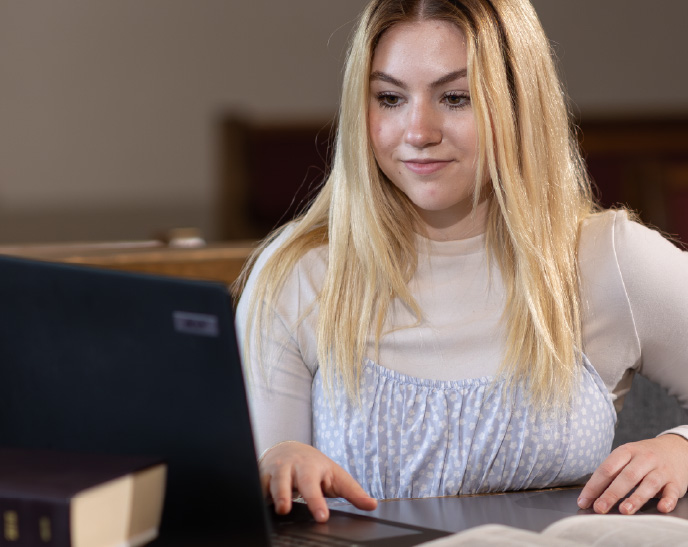 A student studies at a table while using a laptop, with open textbooks nearby. The student looks at the screen with a focused expression in a quiet academic setting.