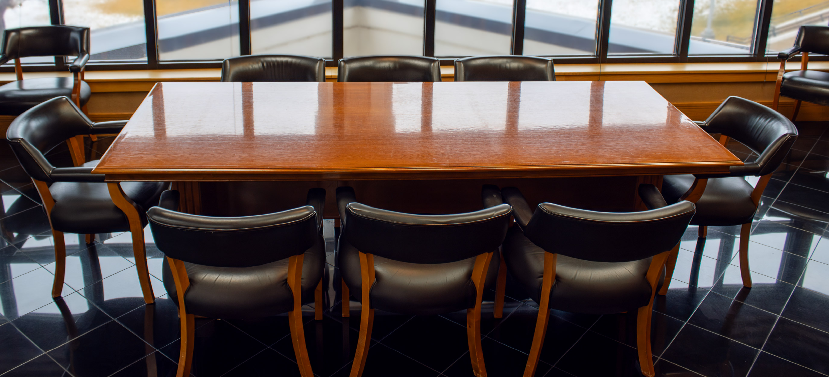 Large polished wooden conference table surrounded by black leather chairs, positioned in front of a wall of windows.