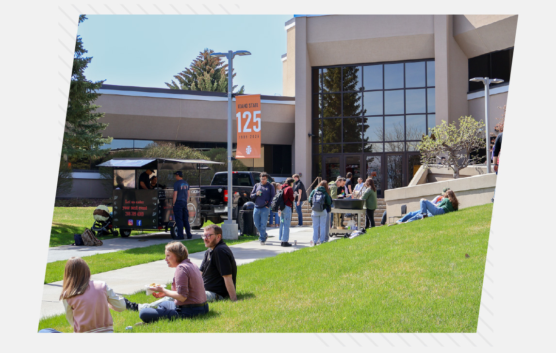 Students gather outside a campus building on a sunny day, eating and socializing on the grass while others visit a food truck and walk along the sidewalk near the entrance.