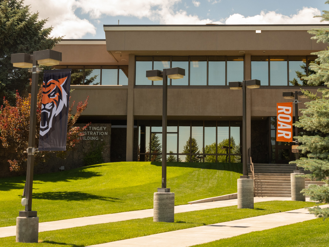 Exterior of the Tingey Administration Building on a sunny day, with campus banners, a walkway leading to the entrance, and landscaped grass and trees in the foreground.