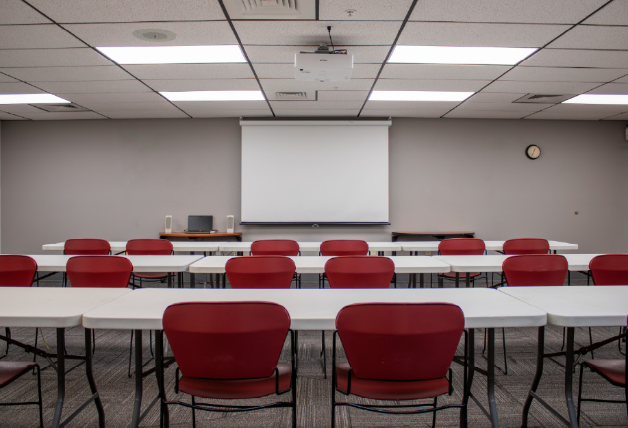 Meeting room arranged in a conference-style layout with white tables forming a rectangle and red chairs around them, facing a projection screen and ceiling-mounted projector, with a laptop and speakers on a side table.