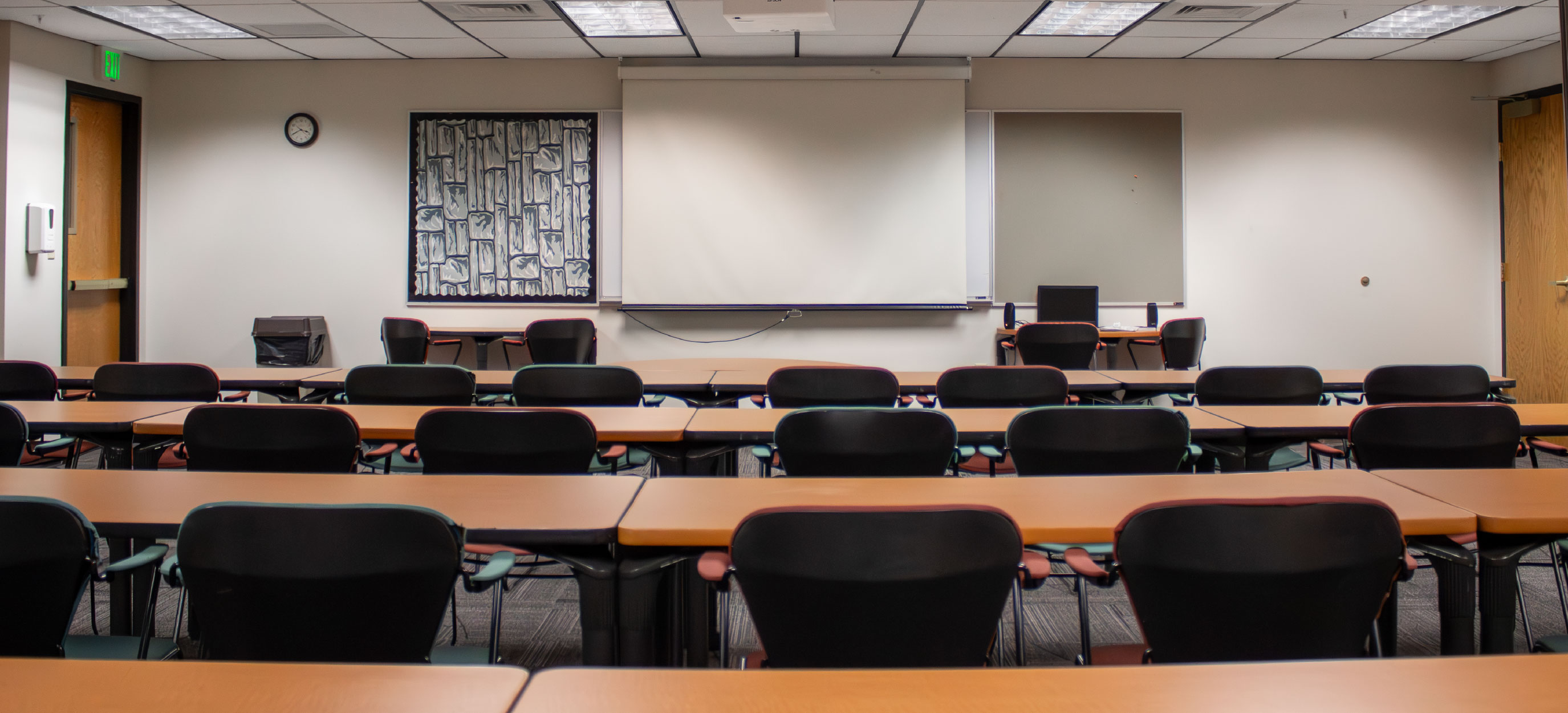 Front view of BSUB 109 showing rows of tables and chairs facing a large projection screen, whiteboard, and instructor workstation.