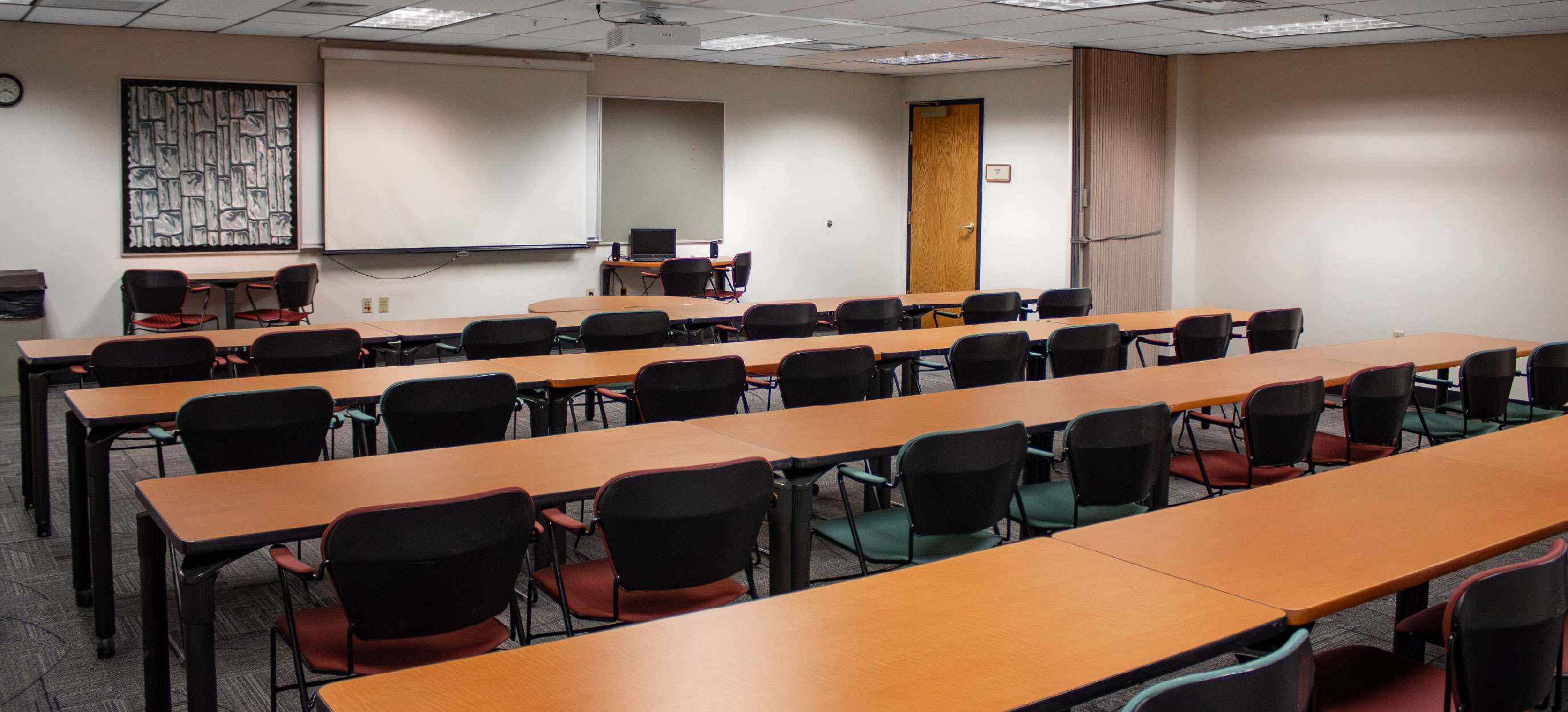 BSUB 109 classroom with rows of long tables and chairs facing a projection screen, whiteboard, and instructor workstation at the front of the room.