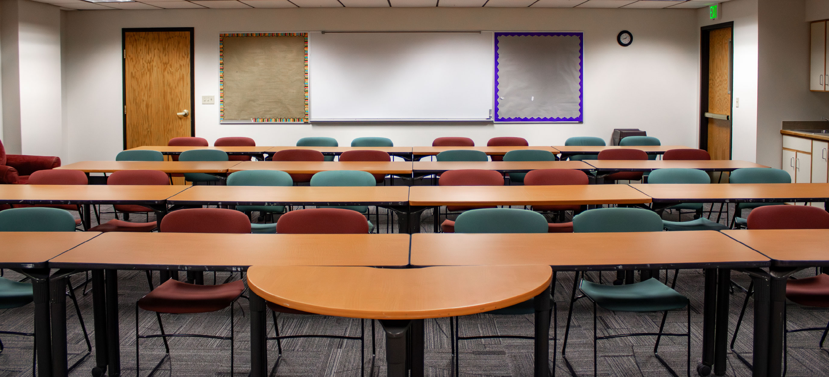 BSUB 109 classroom with rows of tables and red and green chairs facing a large whiteboard, with bulletin boards, a sink area, and doors along the back wall.
