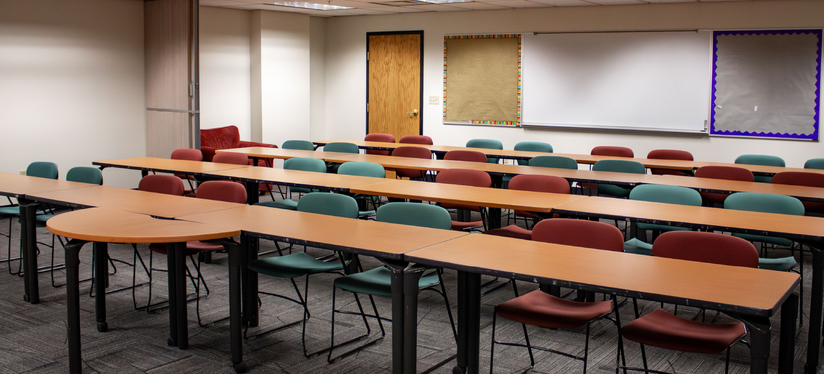 BSUB 109 classroom with long tables and red and green chairs arranged in rows, with a whiteboard and bulletin boards along the back wall.