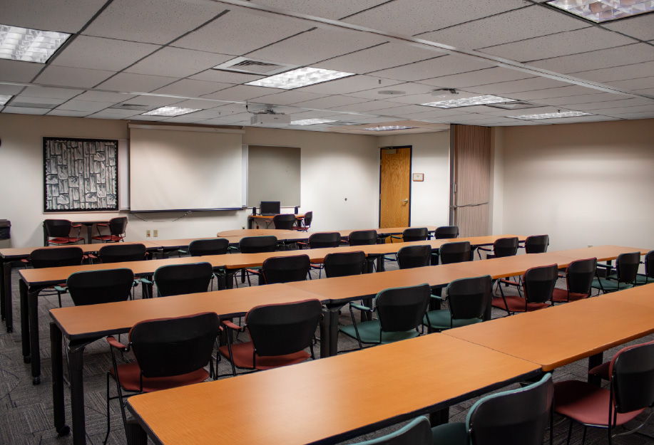 Classroom with rows of long rectangular tables and chairs arranged to face the front of the room, where a projection screen and instructor workstation are located.