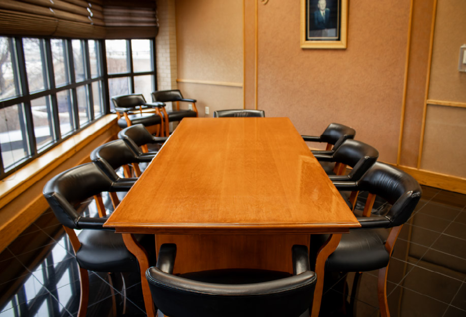 Conference room with a long rectangular wooden table surrounded by black leather chairs. Large windows line one side of the room, with wood blinds and natural light, while warm tan walls and wood trim create a traditional meeting space.