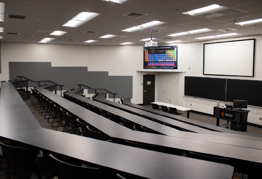 Large tiered classroom with curved rows of desks and chairs facing an instructor station, blackboards, and a large projection screen. A ceiling-mounted projector and periodic table display are visible at the front of the room.