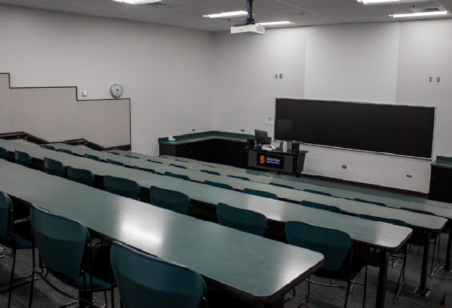 A small tiered classroom with rows of long tables and teal chairs facing the front of the room. The space features a large chalkboard, ceiling-mounted projector, instructor station, and built-in countertops along the front and side walls.