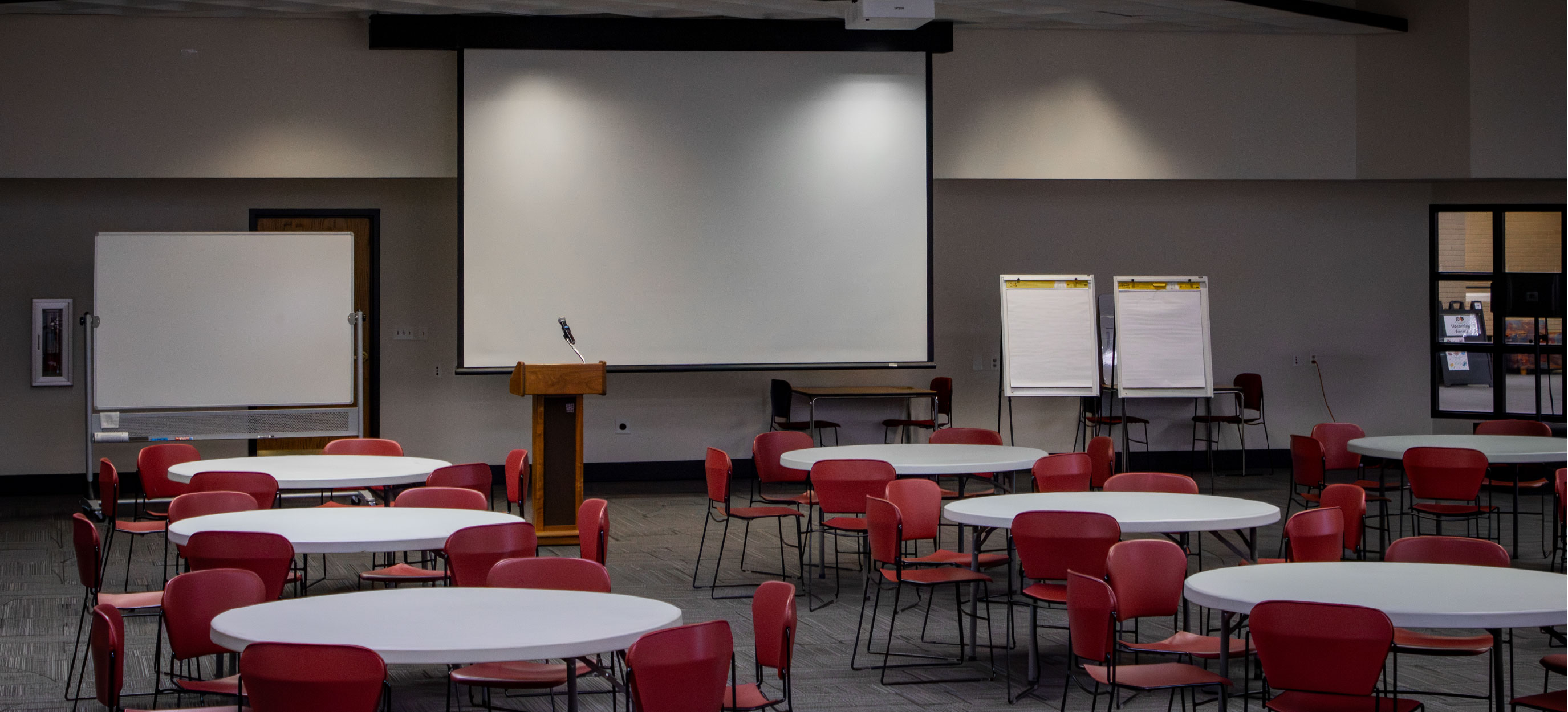 Multipurpose event room arranged with round tables and red chairs, facing a large projection screen, podium, whiteboard, and two flip charts at the front of the space.