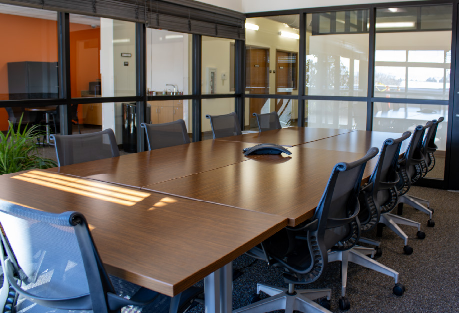 Modern conference room with a large rectangular wooden table surrounded by black mesh office chairs. Glass walls enclose the room, allowing natural light into the space, and a conference phone sits at the center of the table.