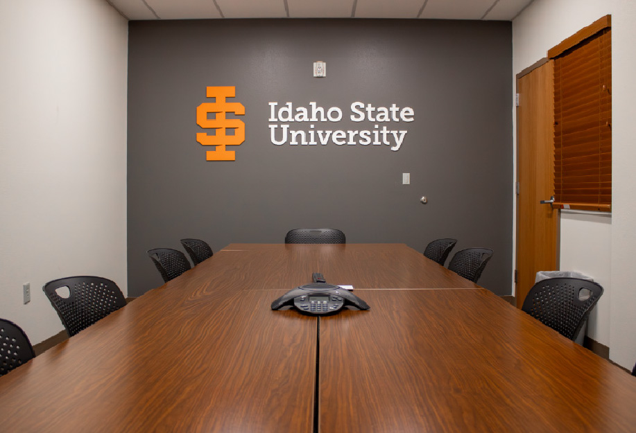 Conference room with a large rectangular wooden table surrounded by black chairs, a conference phone at the center, and an Idaho State University logo displayed on a dark gray accent wall.