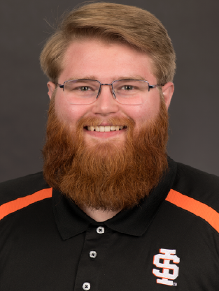 Professional headshot of Noah Mendenhall, transfer coordinator at Idaho State University Idaho Falls. He is smiling and wearing glasses, with short light brown hair and a full red beard. He is dressed in a black Idaho State University polo shirt with orange accents and an ISU logo on the chest, posed against a dark neutral background.