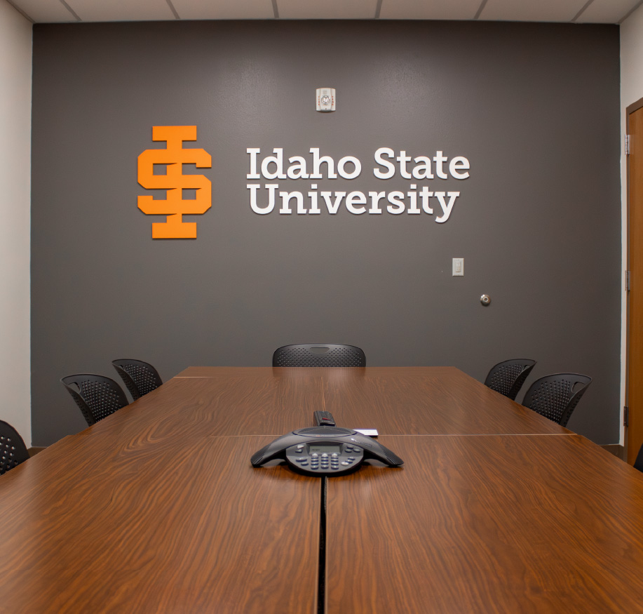 Conference room with a long wooden meeting table, chairs, and a speakerphone, with the Idaho State University logo and name displayed on the wall behind it.