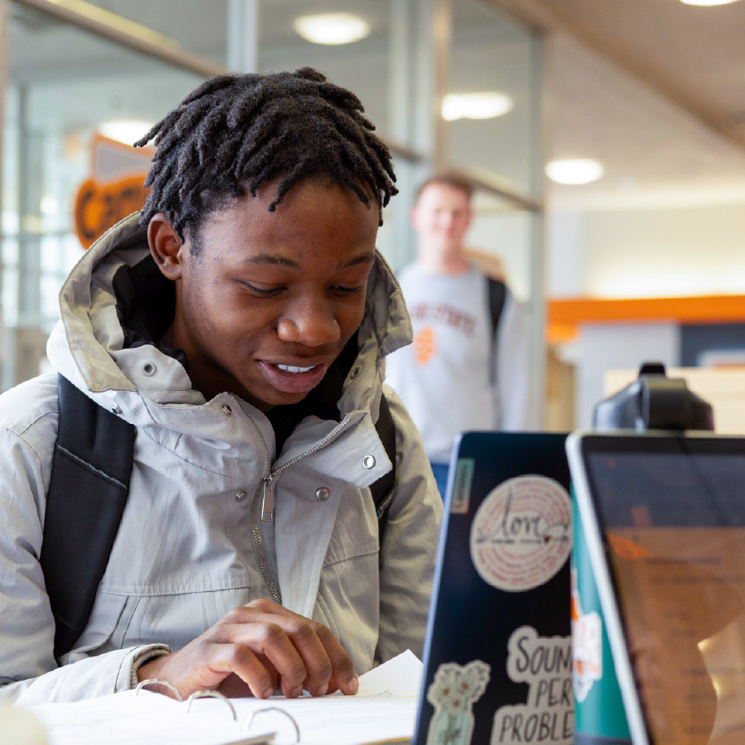 Student with a backpack looking down at papers beside a laptop in a campus study area.