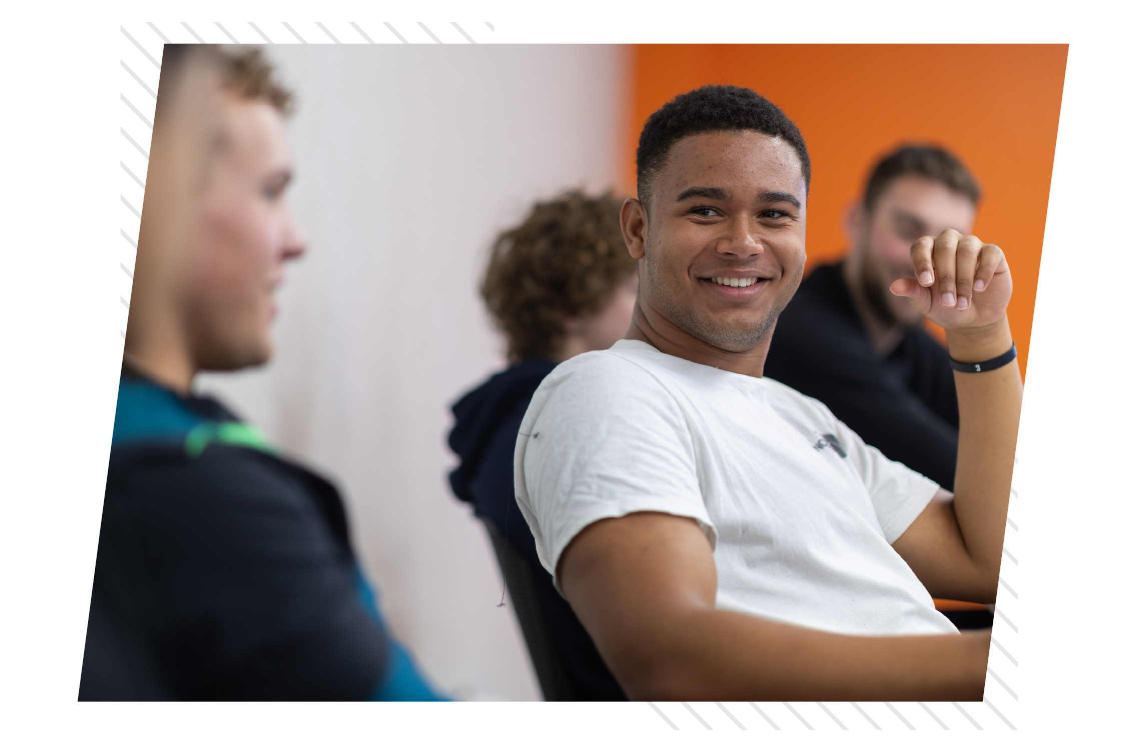 A college student smiles and looks toward a classmate during a classroom discussion. Other students sit nearby in a modern learning space with an orange accent wall, creating an engaged and collaborative academic environment.