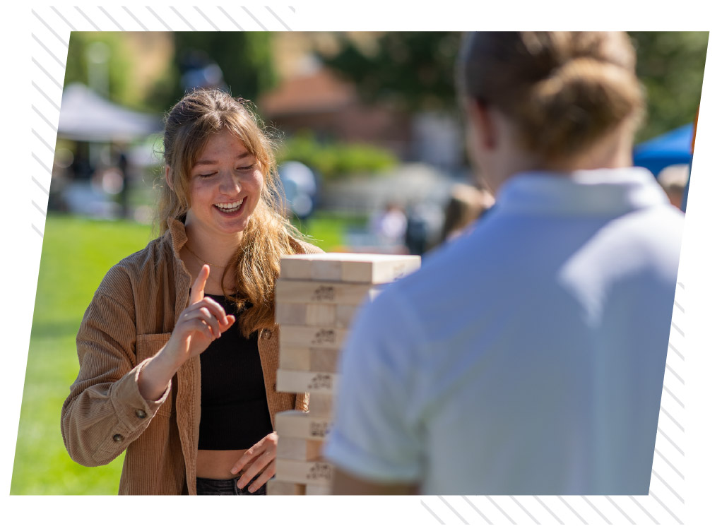 Two students play a giant wooden block-stacking game outdoors during a campus event.