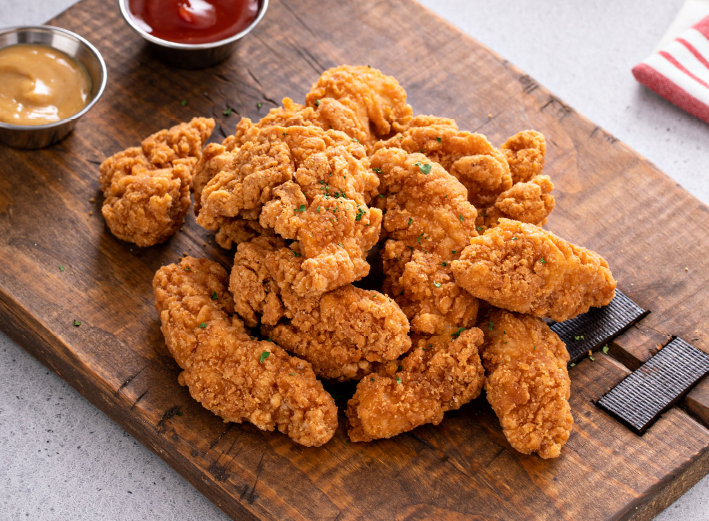 Crispy fried chicken tenders piled on a wooden serving board with dipping sauces in the background.