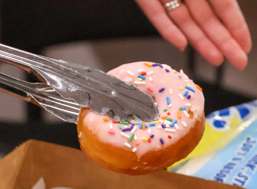Close-up of a hand using metal tongs to pick up a pink-frosted doughnut topped with colorful sprinkles.