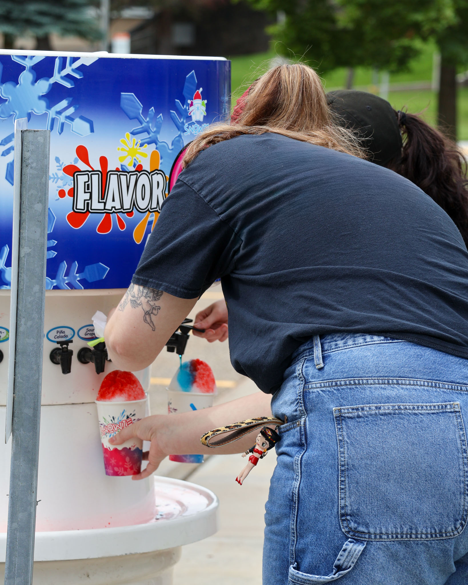 Two people prepare colorful shaved ice at an outdoor snow cone machine, with one person holding a cup of red shaved ice while adding flavoring.
