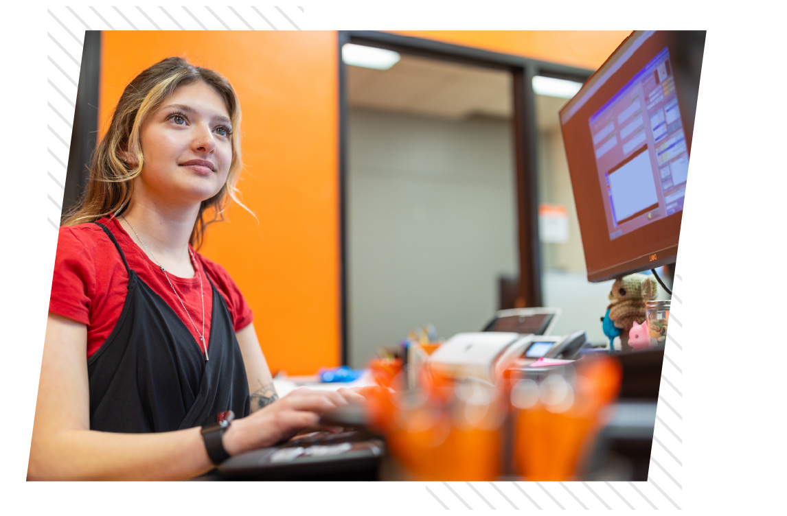 ISU student employee working at a computer workstation, with an orange wall and office equipment in the background.