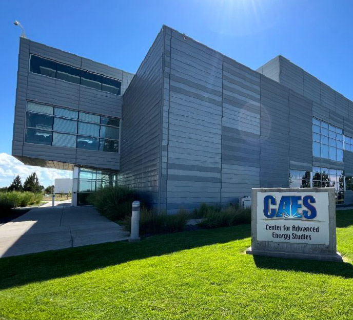 Exterior of the Critical Materials and Energy Systems Innovation Center (CMESIC) at Idaho State University Idaho Falls, featuring a modern gray building with large windows and an entrance walkway, with a sign reading “CAES – Center for Advanced Energy Studies” on the lawn in front.