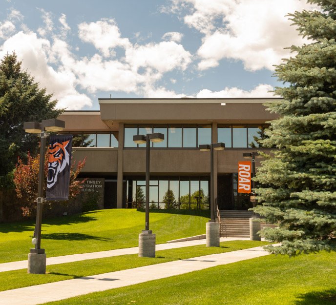 Exterior of the Tingey Administration Building (TAB) at Idaho State University Idaho Falls, featuring a two-story facade with large windows, a central entrance with stairs, landscaped lawns and trees, and campus banners along the walkway.